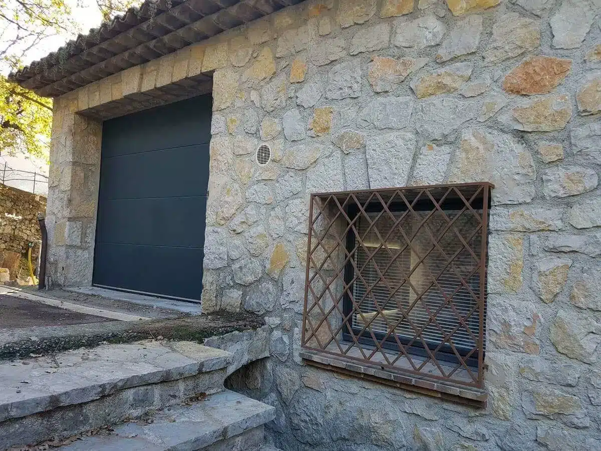 Porte de garage sectionnelle motorisée RAL 7016 installée à Saint-Jeannet, alliant esthétisme contemporain, confort d’utilisation et sécurité renforcée pour une maison en pierre.